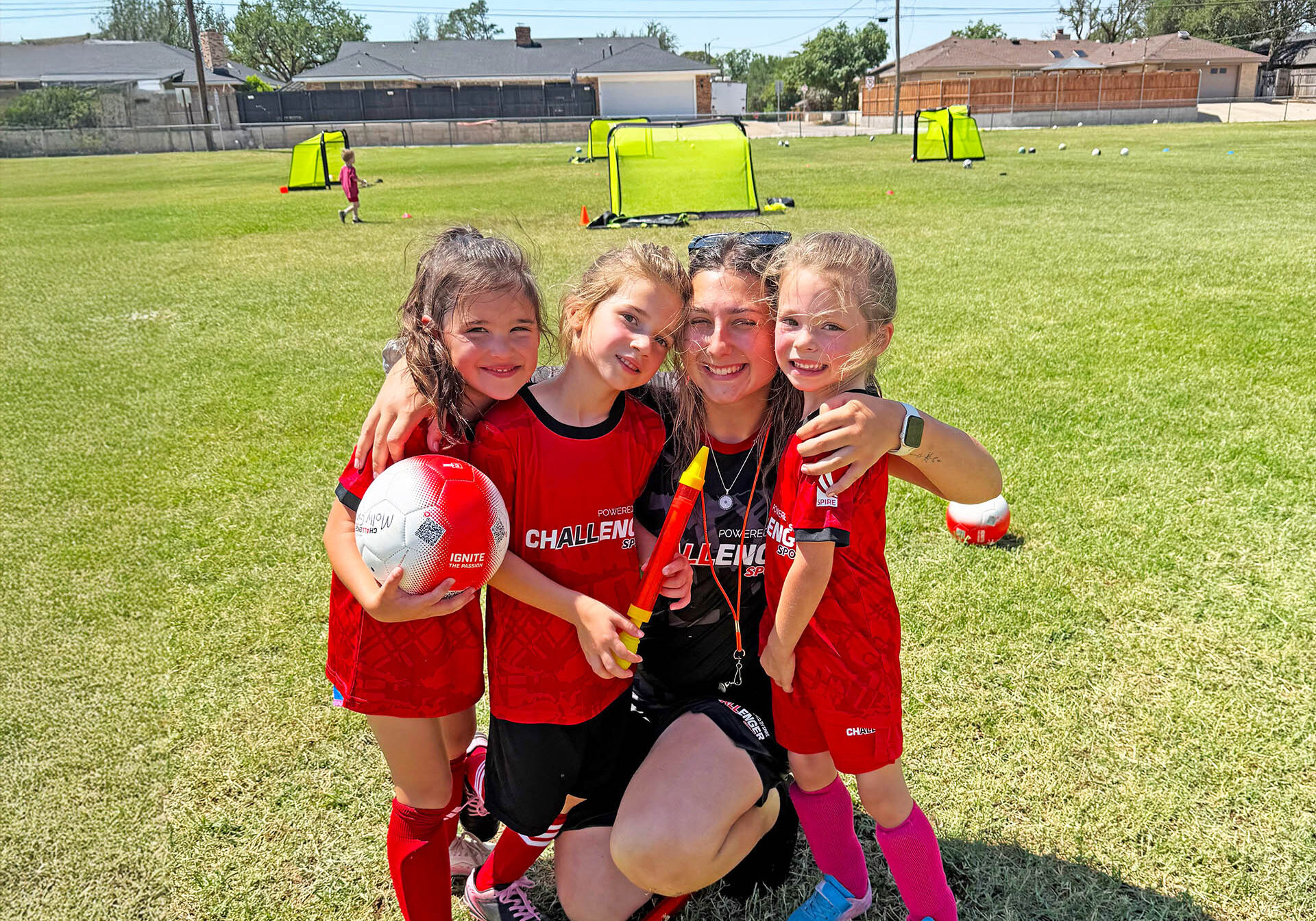 1 female coach kneeling next to 3 female campers arms around them 1 holding soccer ball all wearing Challenger shirts on soccer field posed