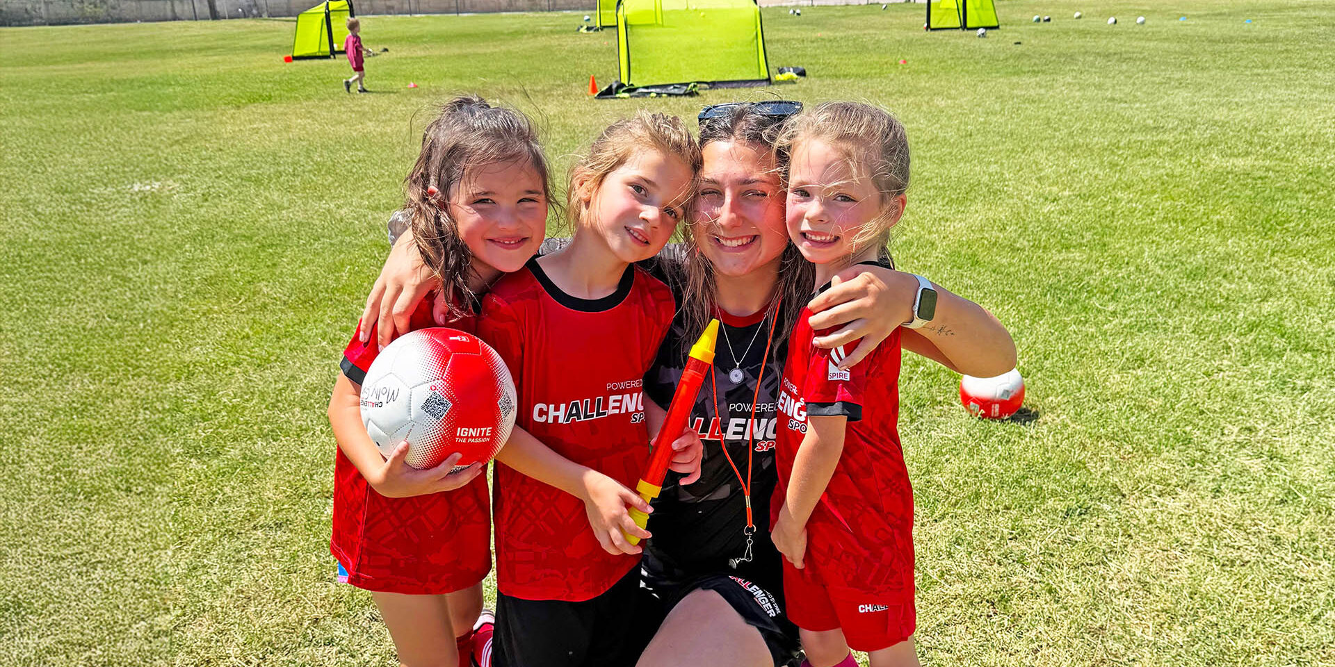 1 female coach kneeling next to 3 female campers arms around them 1 holding soccer ball all wearing Challenger shirts on soccer field posed